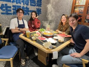 A photo of Abzal and 3 friends, enjoying hot pot. There is steam from rising from the soup bowl and everyone is smiling at the camera for the photo.